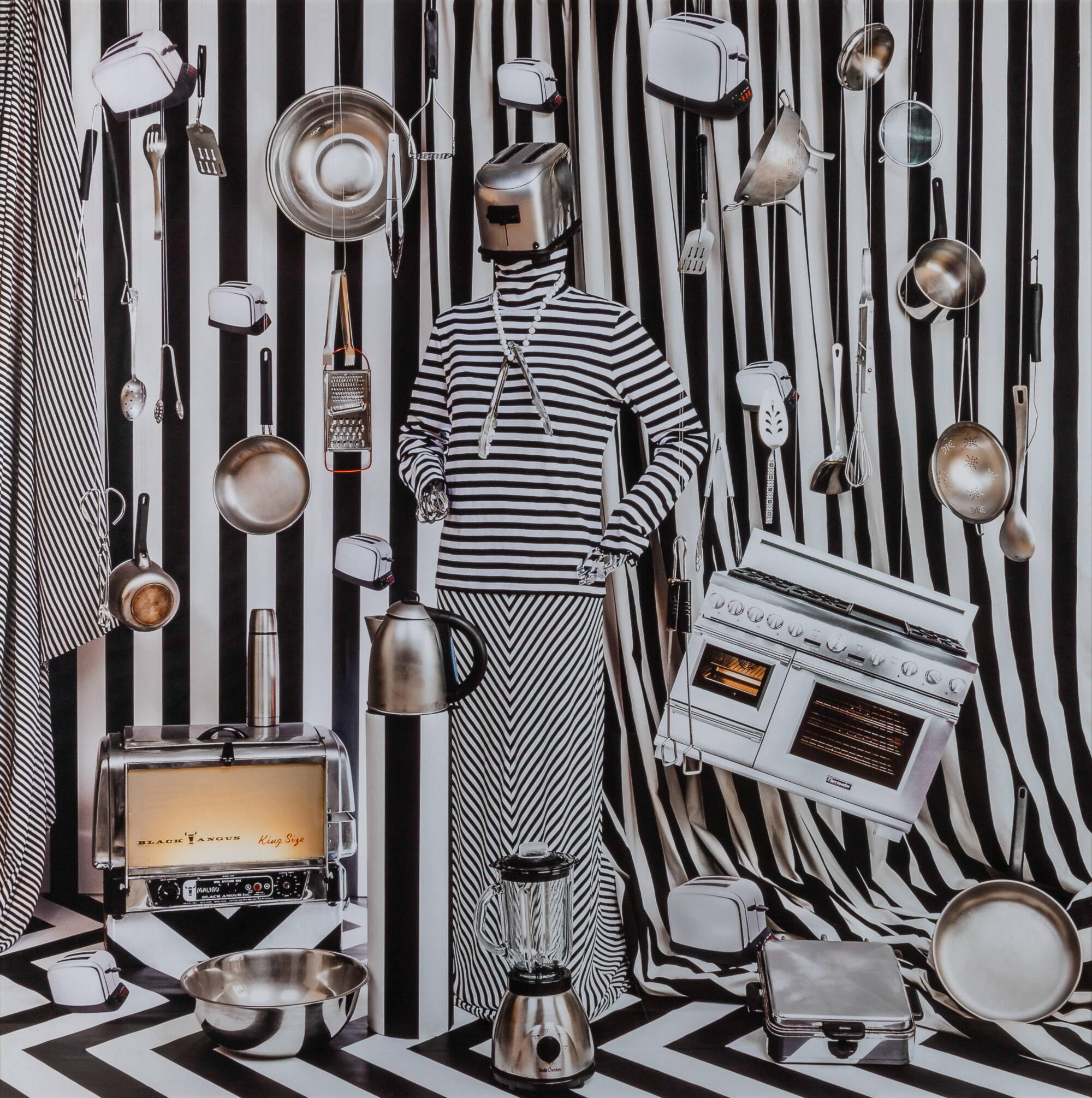 Photograph of a female form wearing black and white stripes with a toaster for a head. Background includes more black and white stripes and a variety of other kitchen appliances including a stove, a blender and pots and pans.